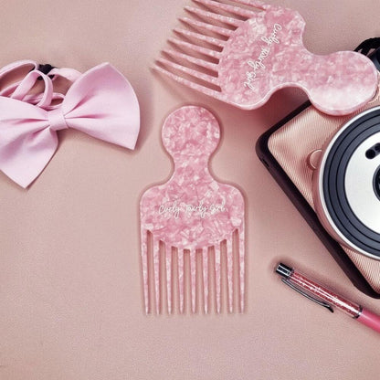 Pink afro hair comb on table with camera, pen and bow